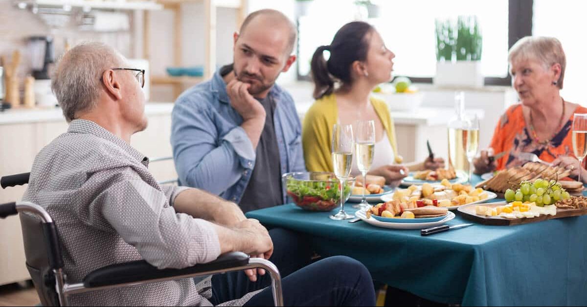 Image of an older man in a wheelchair is talking to his son while the family is sitting around the dinner table