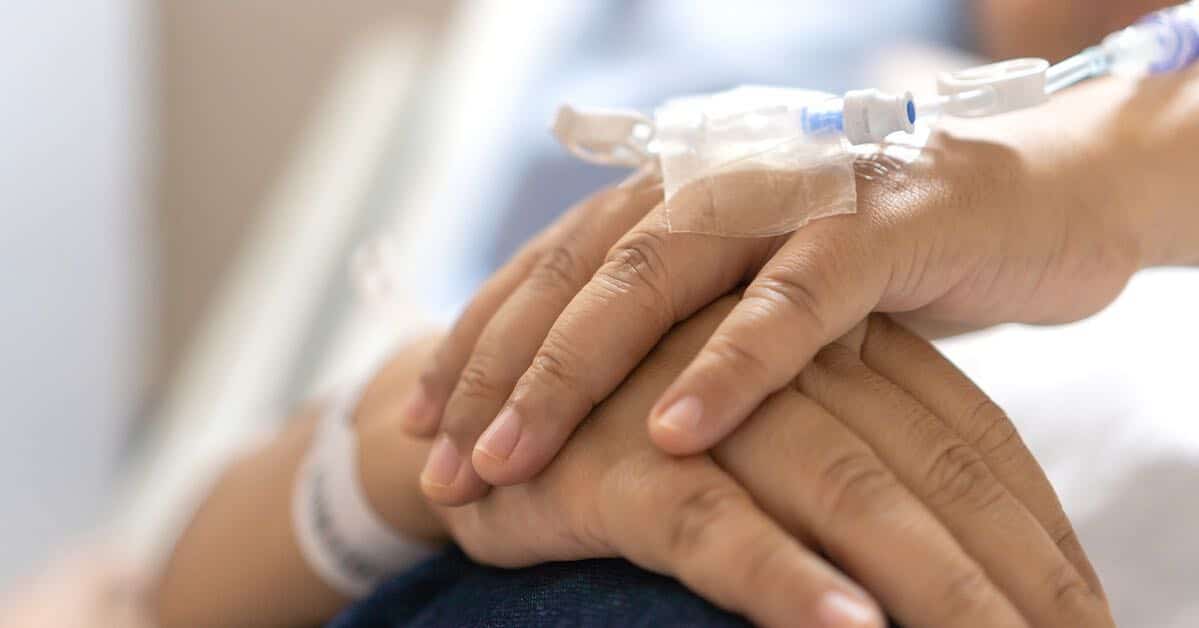 An elderly patient in bed with an IV in his hand, close up on him holding hands with a loved one