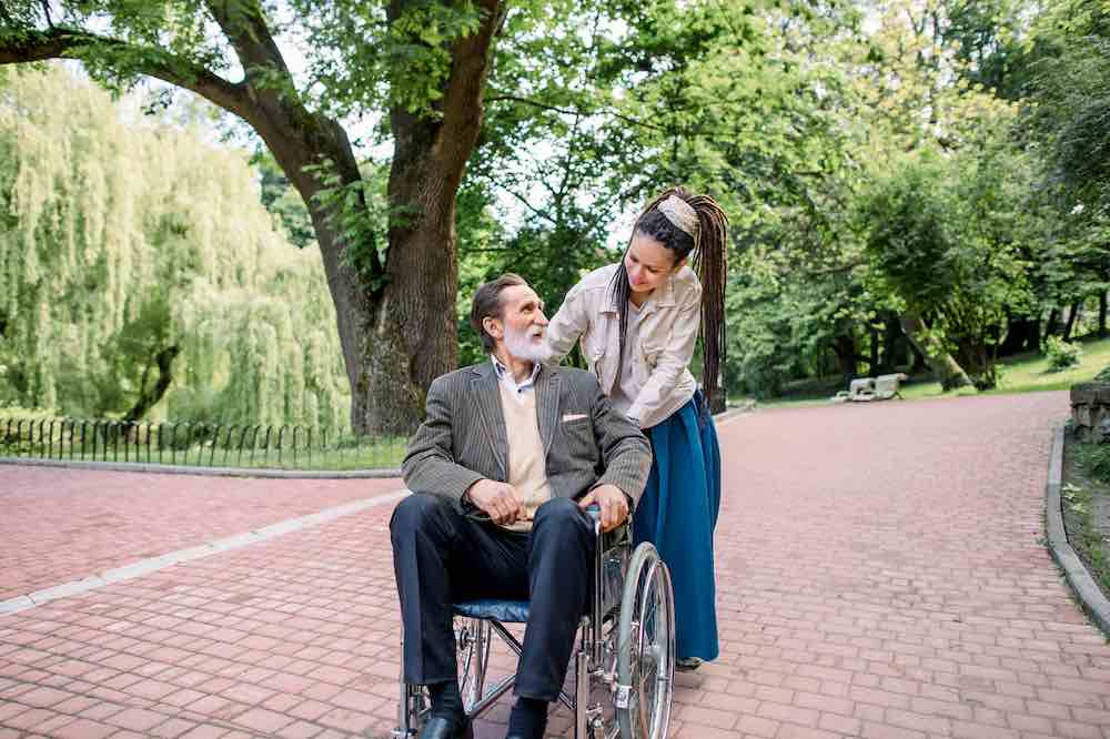 Disabled-senior-bearded-man-in-wheelchair-and-his-creative-girl-assistant-with-dreadlocks-