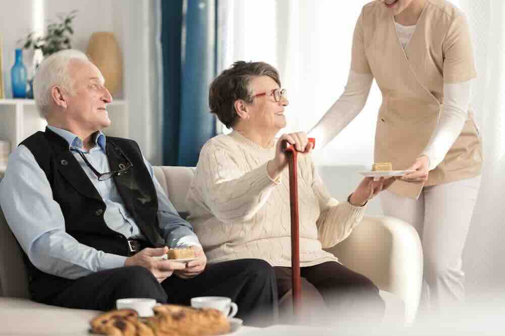 Elderly couple sitting on a couch being helped by an in home hospice caregiver.