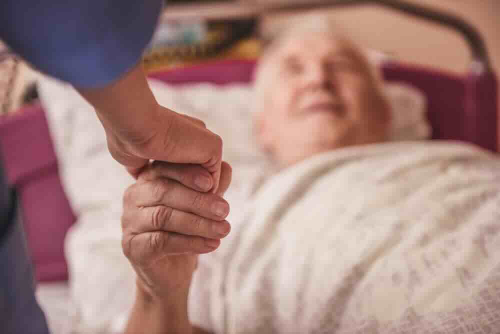 Selective focus of an elderly male patient on a bed holding the hand of an in home hospice caregiver.