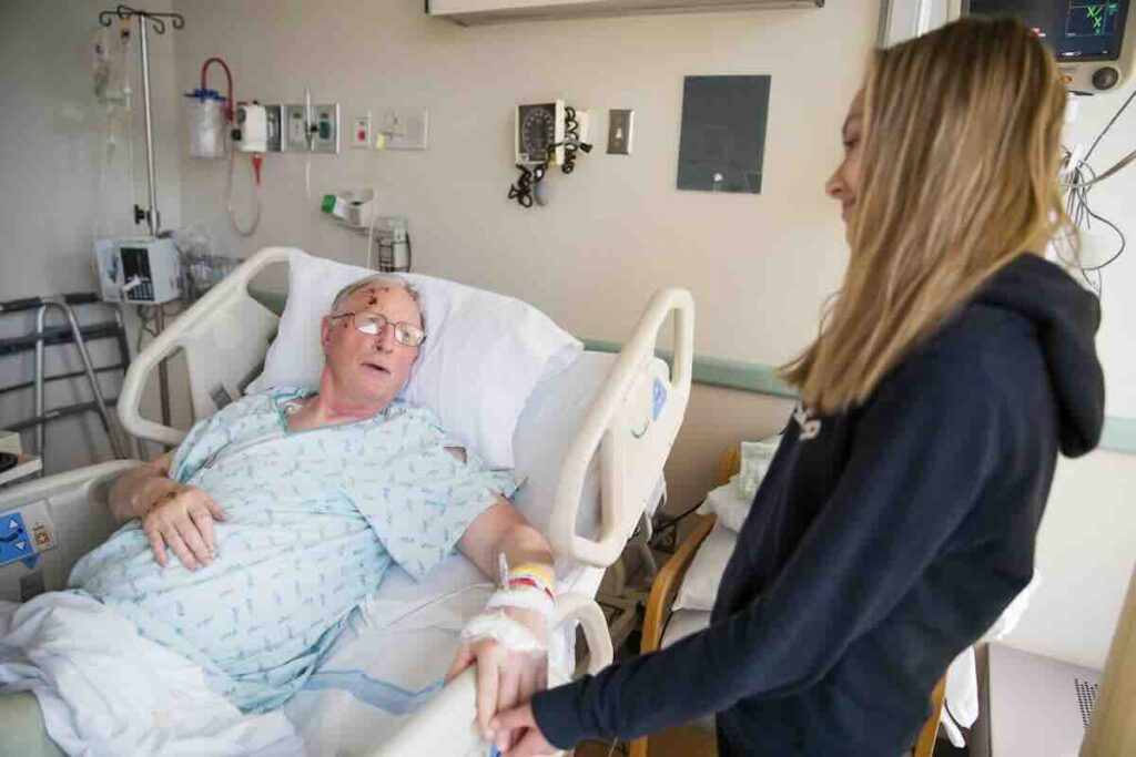 Senior man on a hospital bed talking with a blonde hospice volunteer.
