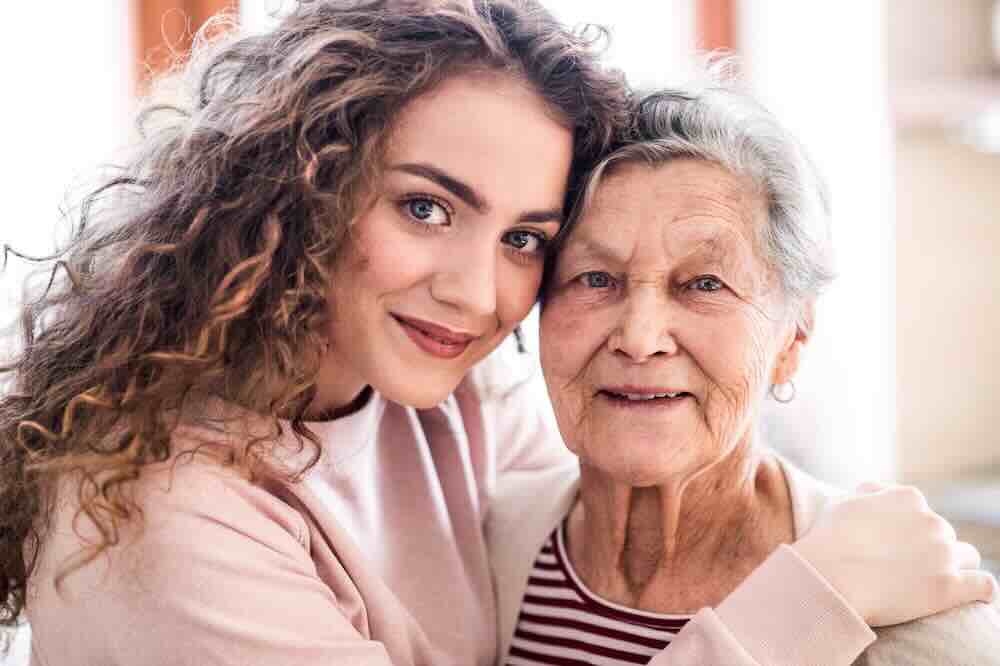 Senior woman hugging a young woman with curly hair who’s her in home hospice caregiver.