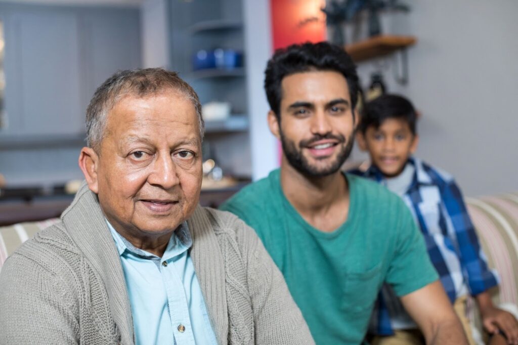 A senior man, his son, and his grandson are seated on the couch asking “What do home health aides do?”